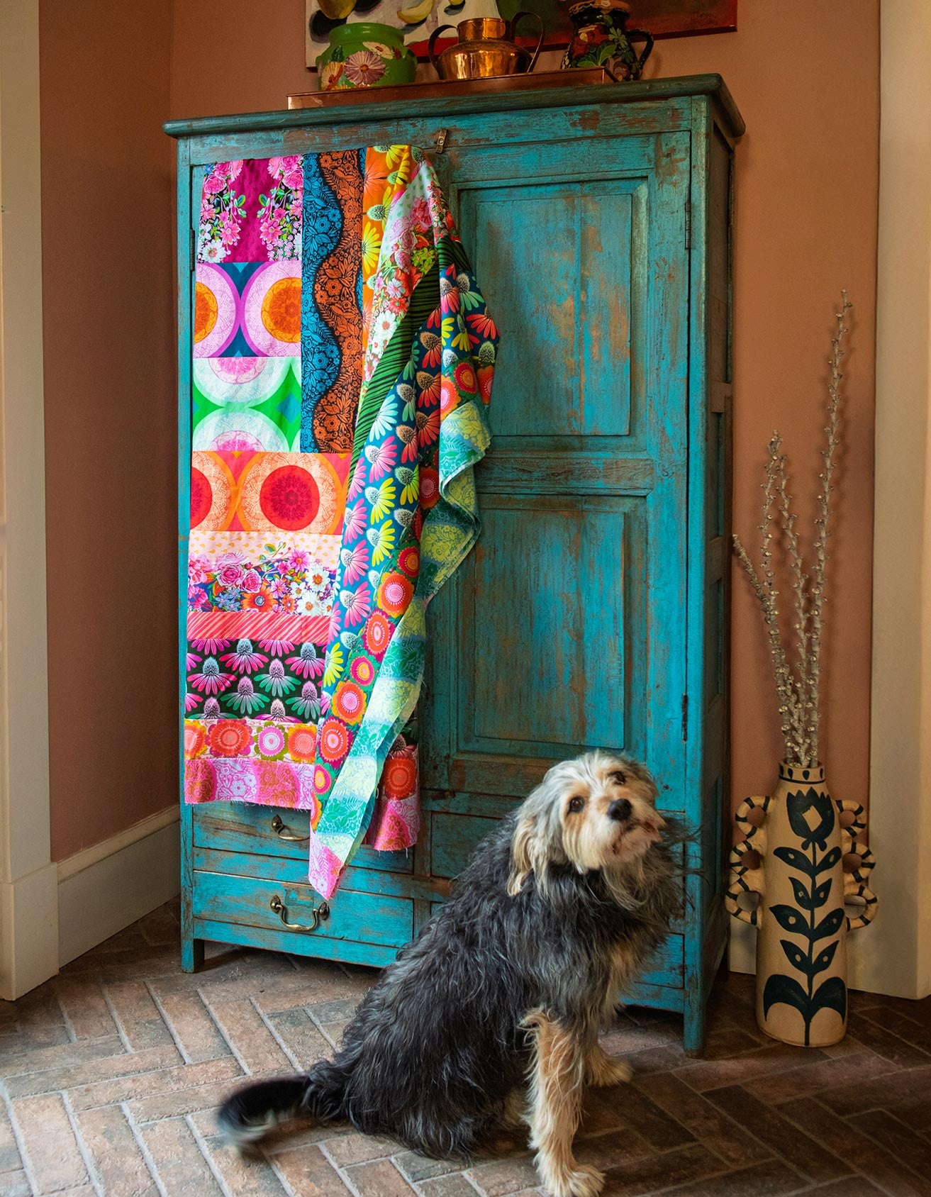 A dog sits in front of a vibrant, colorful quilt hanging on a blue wooden cabinet.