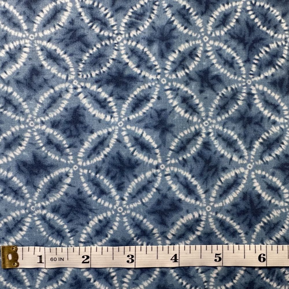 a close-up view of a fabric with a repeating pattern of blue and white geometric shapes, resembling a tie-dye design. A ruler is placed in front of the fabric to provide a scale reference.