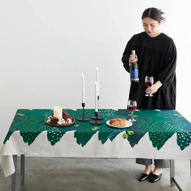 A woman in a black dress stands at a table with a green tablecloth, holding a bottle of wine and a glass, while a plate of food and a candle holder are also present on the table.
