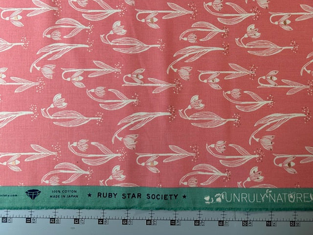 a fabric with a repeating pattern of white flowers on a pink background. The fabric appears to be made of cotton and is displayed on a ruler for size reference.