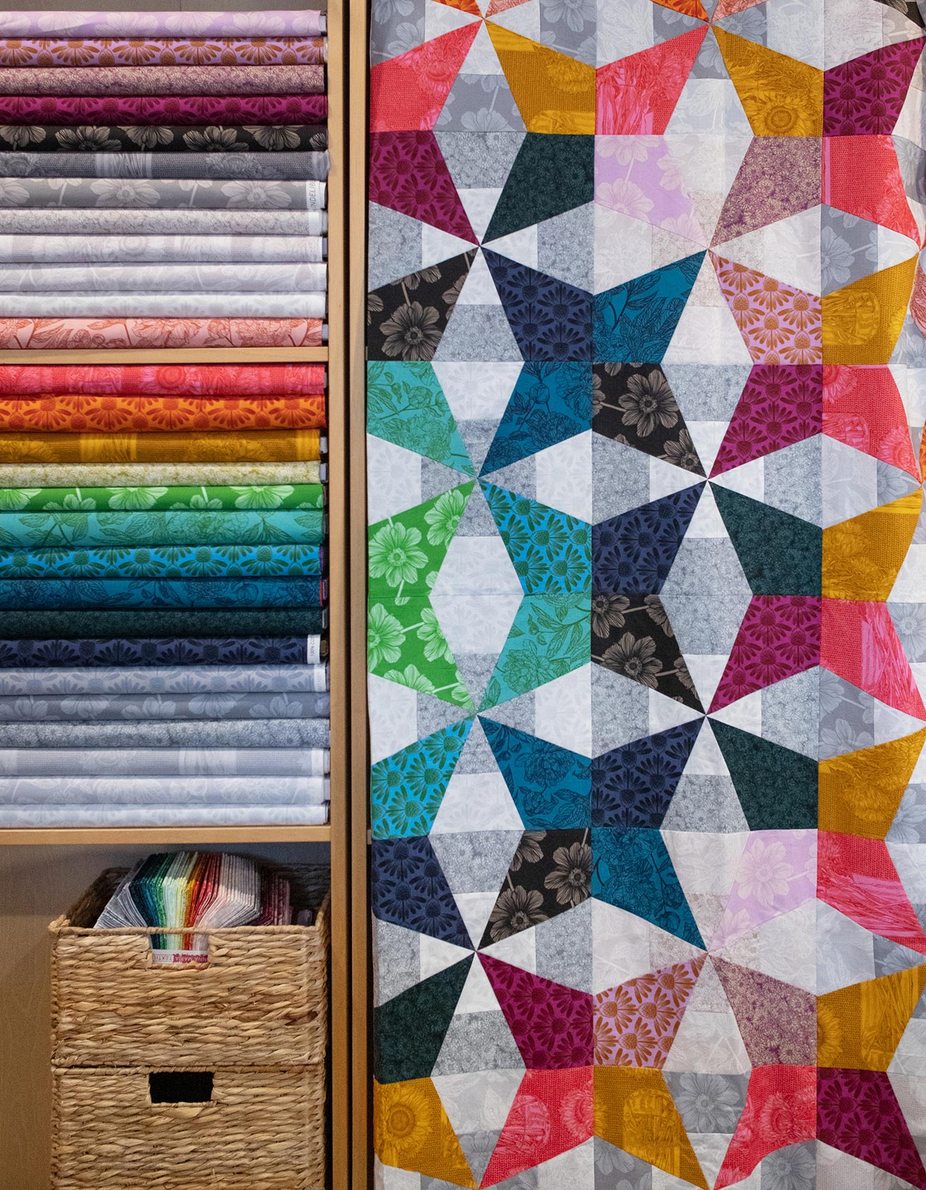 a vibrant and colorful quilt with a geometric pattern of triangles and squares in various shades of pink, blue, green, and yellow. The quilt is displayed next to a wooden shelf filled with fabric rolls, creating a visually appealing and organized display.
