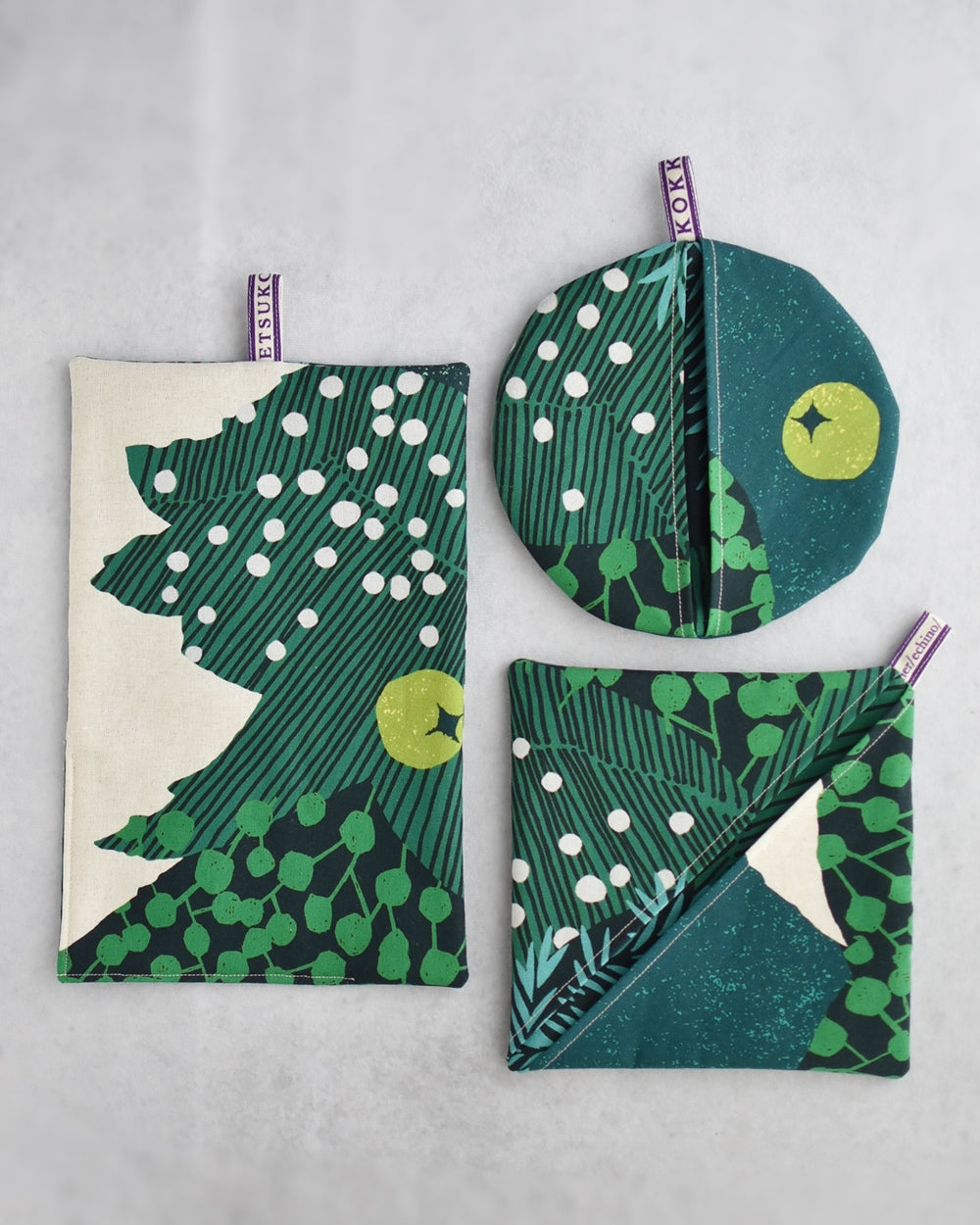 three green and white patterned fabric items, including a rectangular towel, a circular pot holder, and a square pouch, all with a similar green and white design.