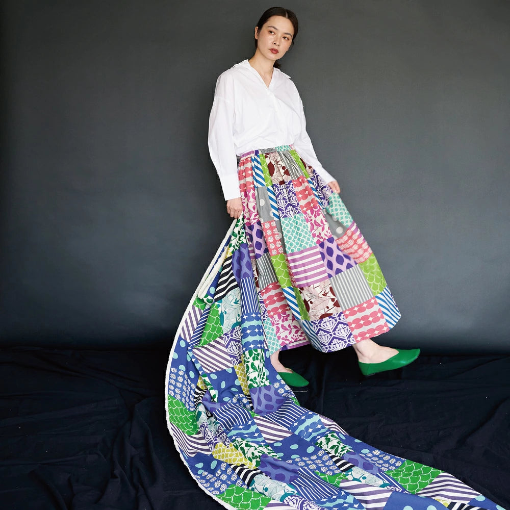 A woman wearing a white blouse and a colorful, patterned skirt stands against a dark background, with a long, flowing skirt trailing behind her.