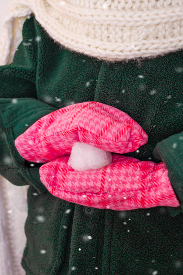 A person wearing a green jacket and pink and white patterned mittens is holding a white ball in their hands, with snow falling around them.