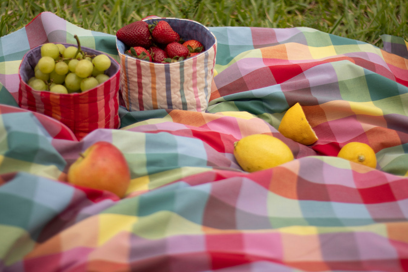 A picnic blanket spread out on a grassy field, with two baskets of fruit - one filled with red strawberries and the other with green grapes. There are also two apples and two lemons placed on the blanket.