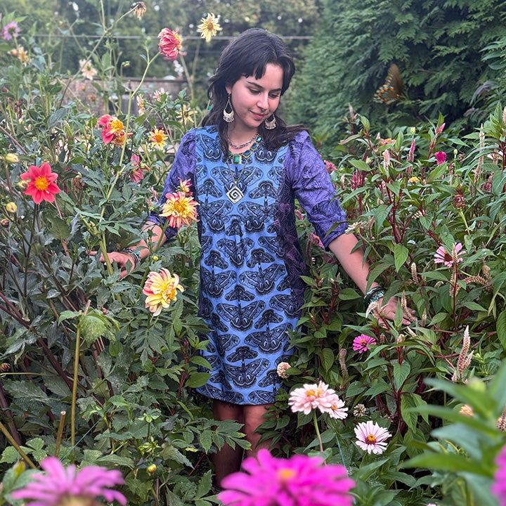A woman in a blue and black patterned dress stands amidst a vibrant garden filled with colorful flowers, including pink, yellow, and orange blooms.