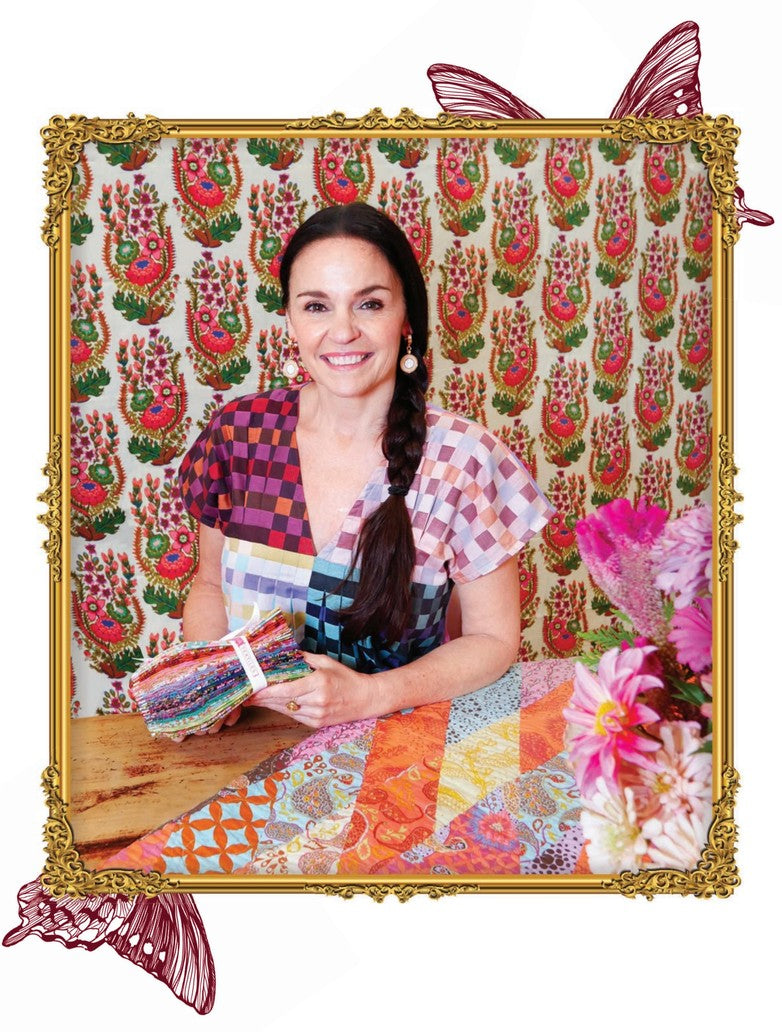 A woman is sitting at a table, holding a bundle of fabric, surrounded by a colorful floral patterned wall and pink flowers.