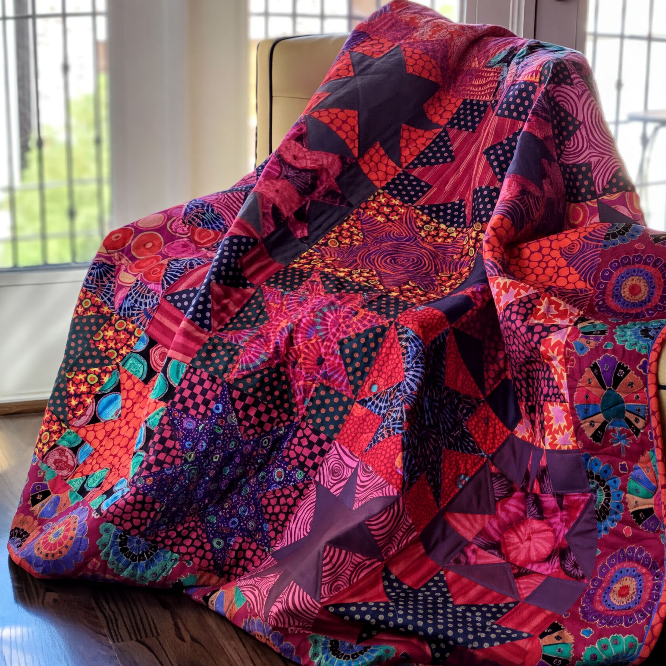 a vibrant, colorful quilt with a variety of patterns and designs, including stars, flowers, and geometric shapes, all in shades of red, purple, and blue. The quilt is displayed on a wooden surface, possibly a table or a chair, and is positioned in front of a window, allowing natural light to illuminate the intricate details of the quilt.