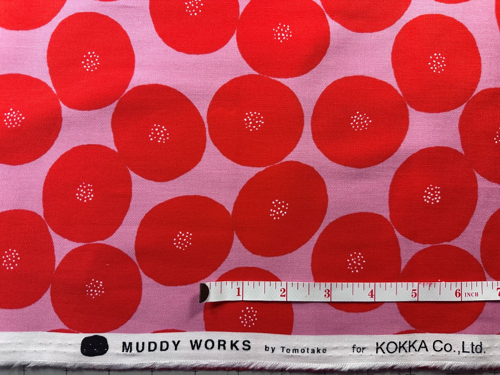a fabric with a repeating pattern of red circles with white spots, creating a vibrant and eye-catching design. The fabric is displayed alongside a ruler for scale, indicating its size.