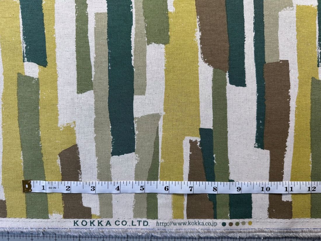a fabric with a striped pattern in various shades of green, yellow, and brown. A ruler is placed in front of the fabric, indicating the size of the fabric.