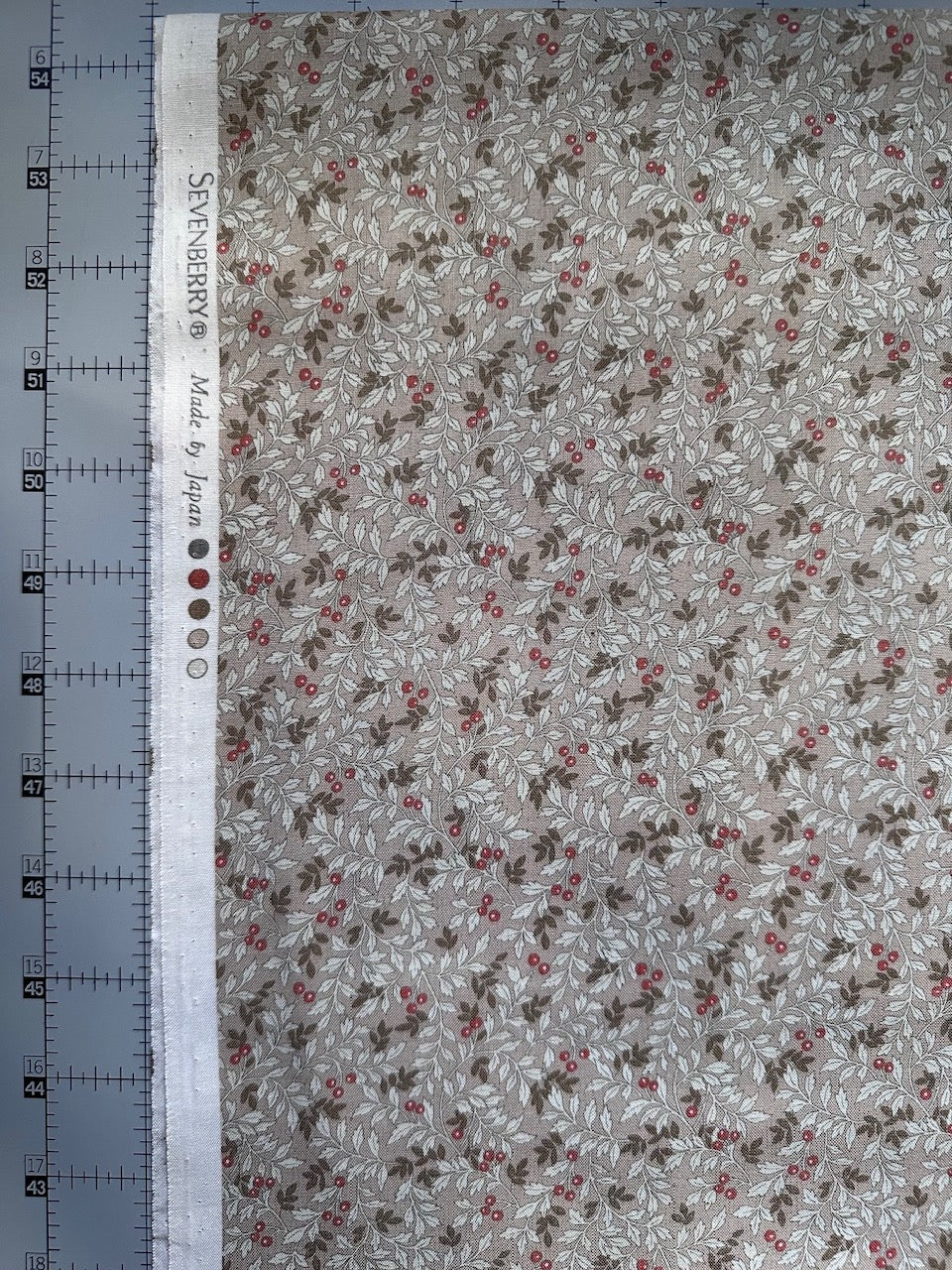 a fabric with a floral pattern, featuring leaves and red berries. The fabric appears to be made of a light-colored material, possibly cotton or linen. There is a ruler placed next to the fabric, which is likely used for measuring the fabric's dimensions.