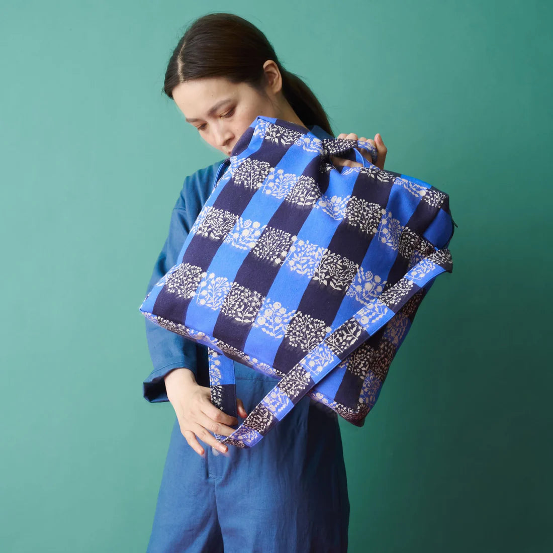 A woman in a blue jumpsuit is holding a large blue and white checkered bag, looking down at it with a thoughtful expression.