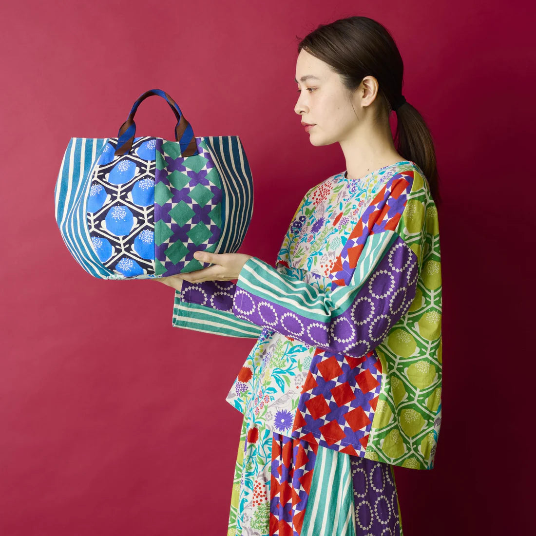 A woman in a colorful, patterned dress is holding a large, patterned bag against a red background.