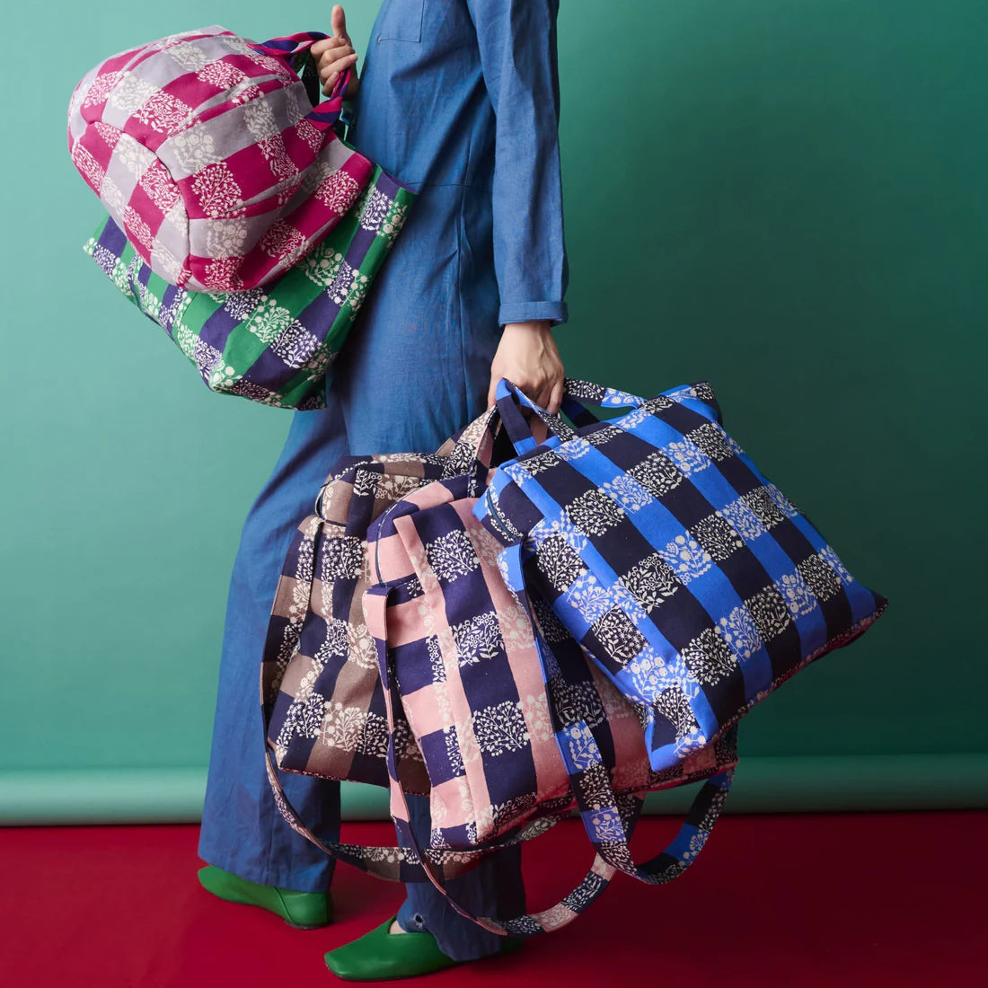 A person is standing against a green background, holding three colorful bags in their hands.
