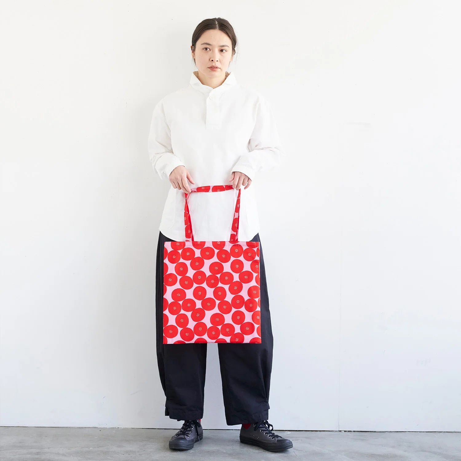 A woman in a white shirt and black pants stands against a white wall, holding a red bag with a red and white polka dot pattern.