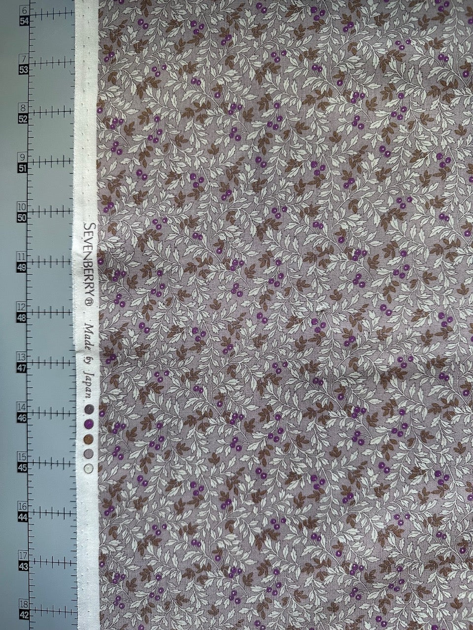 a fabric with a floral pattern, featuring purple and brown flowers and leaves. The fabric is displayed on a ruler for scale, indicating its size.