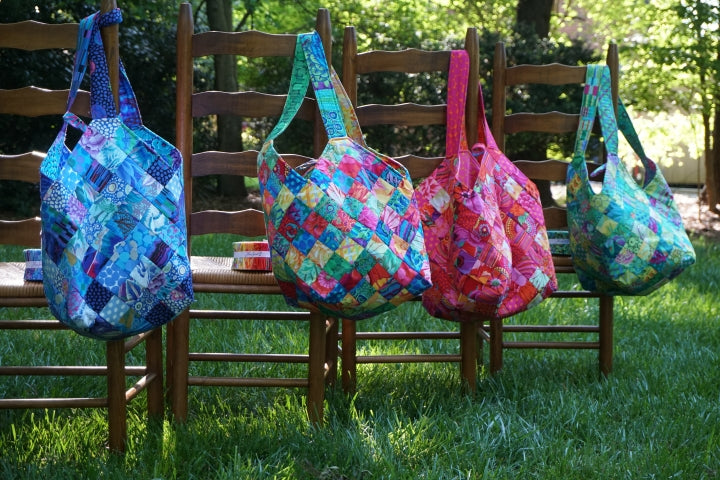 Four colorful quilt bags are hanging on wooden chairs in a grassy outdoor setting.