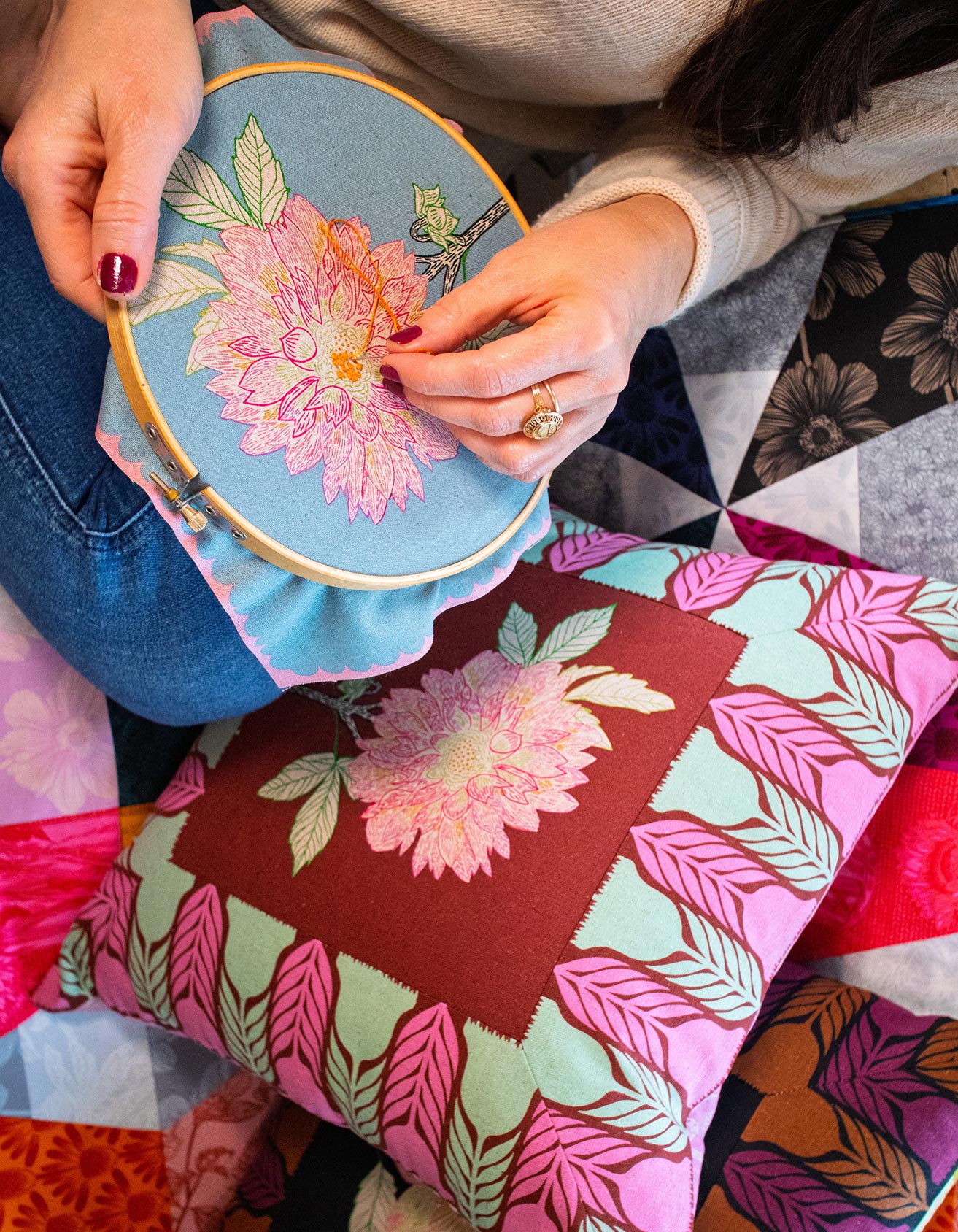 A person's hands are holding a blue embroidery hoop with a pink flower design, and they are working on a colorful quilt or pillow with a floral pattern.