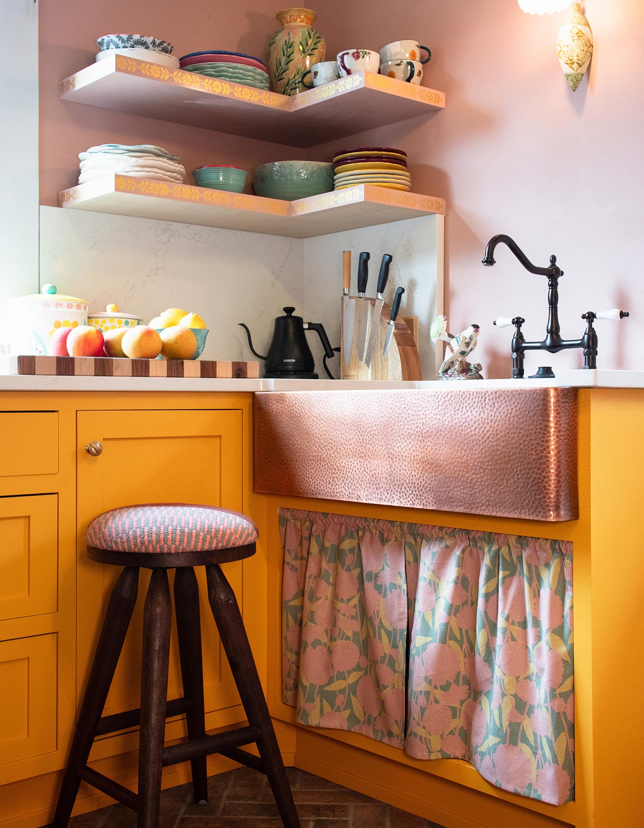 a kitchen with a yellow cabinet, a black sink, and a wooden stool. The countertop has various kitchen items such as a kettle, a knife set, and a bowl. The walls are painted pink, and there are shelves above the sink holding decorative items.