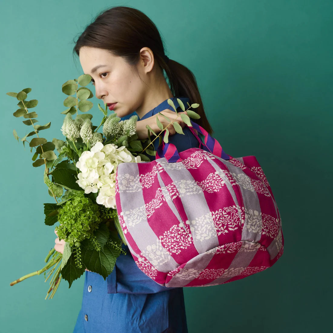 A woman in a blue dress holds a large bouquet of flowers and a pink and white checkered bag, standing against a teal background.