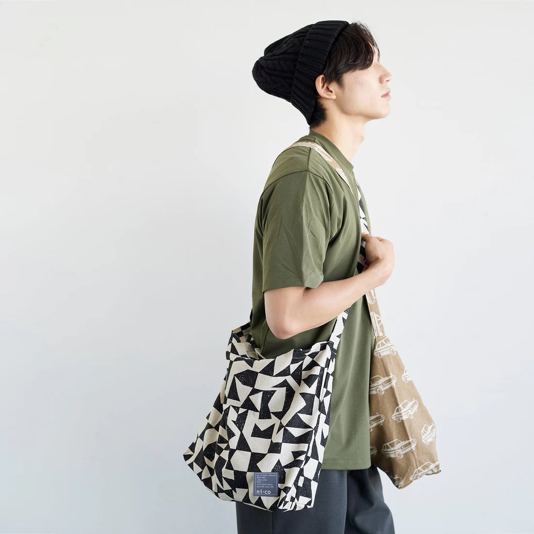 A person wearing a green shirt and a black beanie is standing against a white background, carrying a patterned tote bag.