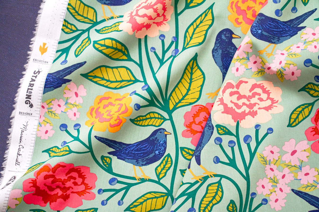 a vibrant and colorful fabric with a repeating pattern of blue birds, flowers, and leaves in shades of pink, yellow, and green.