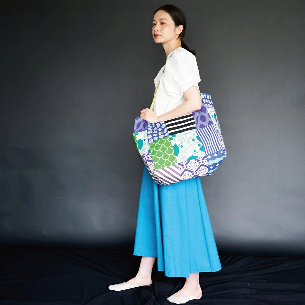 A woman wearing a white top and blue skirt stands against a black background, holding a colorful bag.