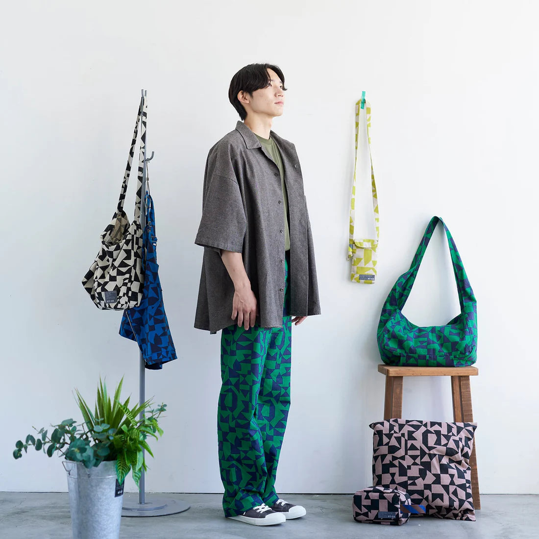 A person wearing a gray jacket and patterned pants stands in a room with various bags and plants.