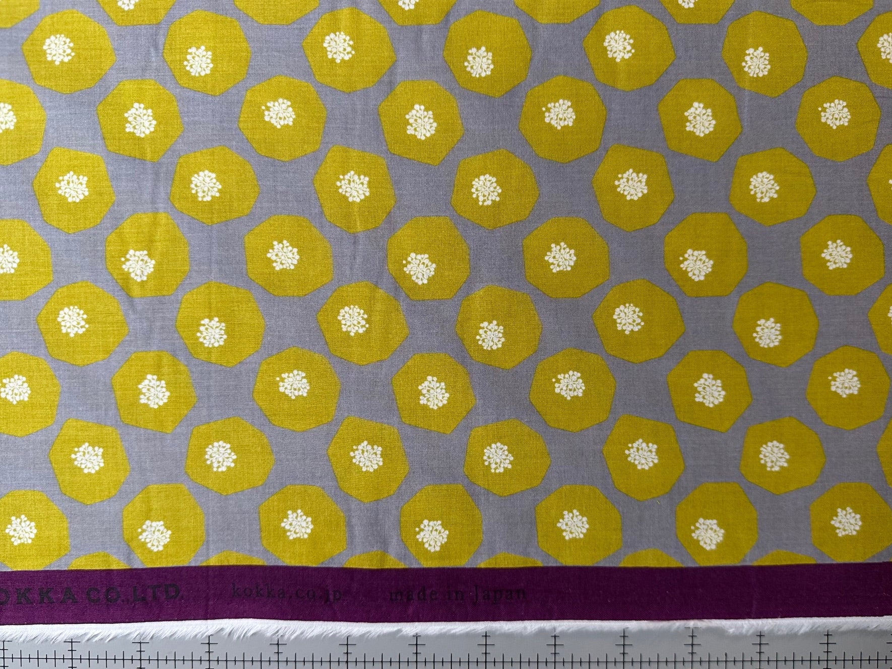 a fabric with a repeating pattern of yellow and gray hexagons, with small white flowers or dots scattered throughout the design. The fabric appears to be a type of quilt or textile, with a purple border running along the bottom edge.