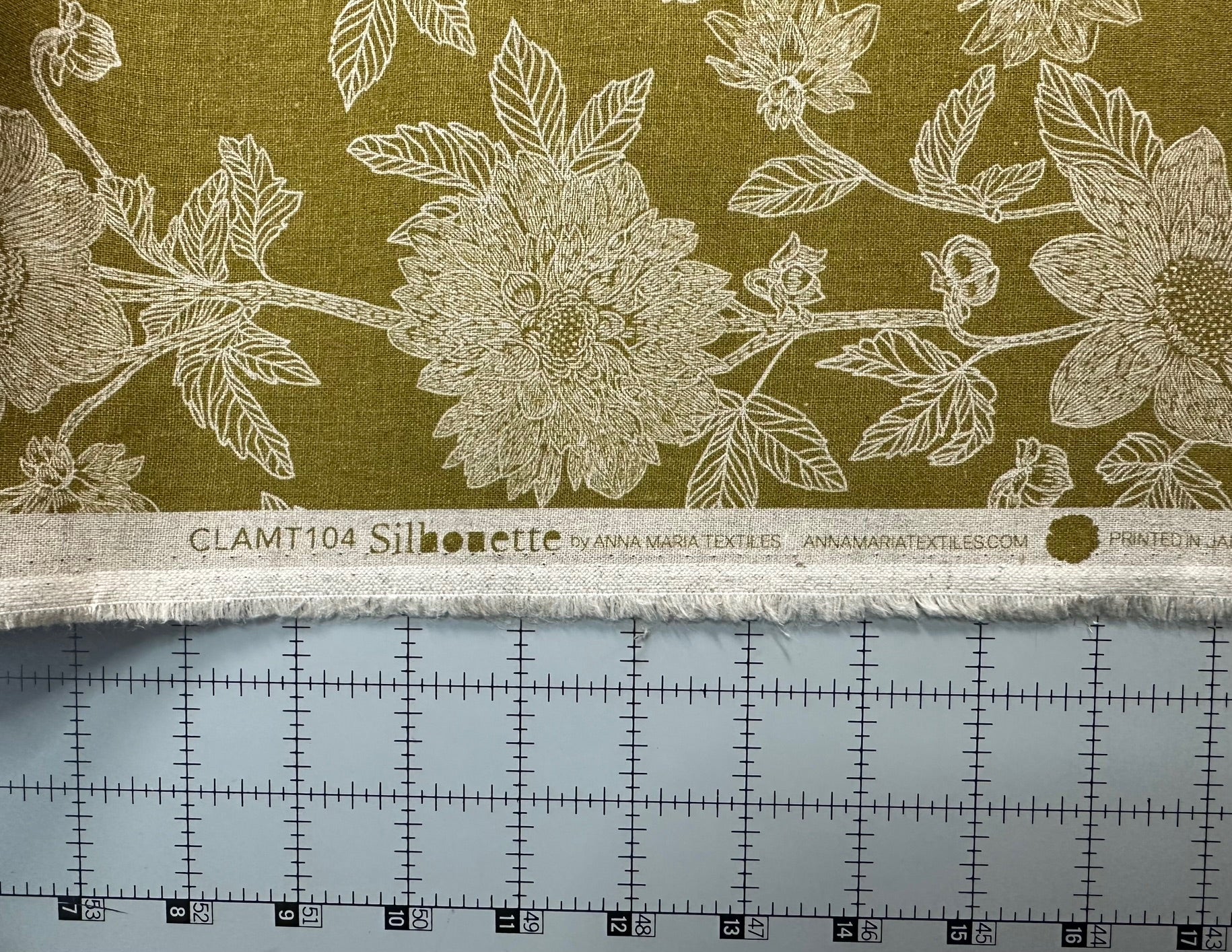 a close-up view of a fabric with a floral pattern, including large white flowers and leaves. The fabric appears to be a type of silhouette material, as indicated by the text "CLAMT104 Silhouette" and "ANNA MARINA TEXTURES" on the fabric. The fabric is displayed on a measuring mat, which is a common tool used in sewing and tailoring to ensure accurate measurements and proper cutting.