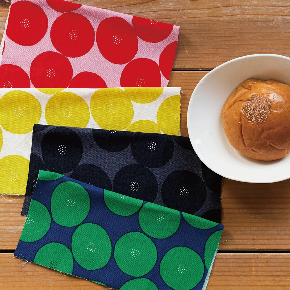 a wooden table with a white bowl containing a sandwich, a red and yellow polka dot cloth, a blue and green polka dot cloth, and a green and white polka dot cloth.