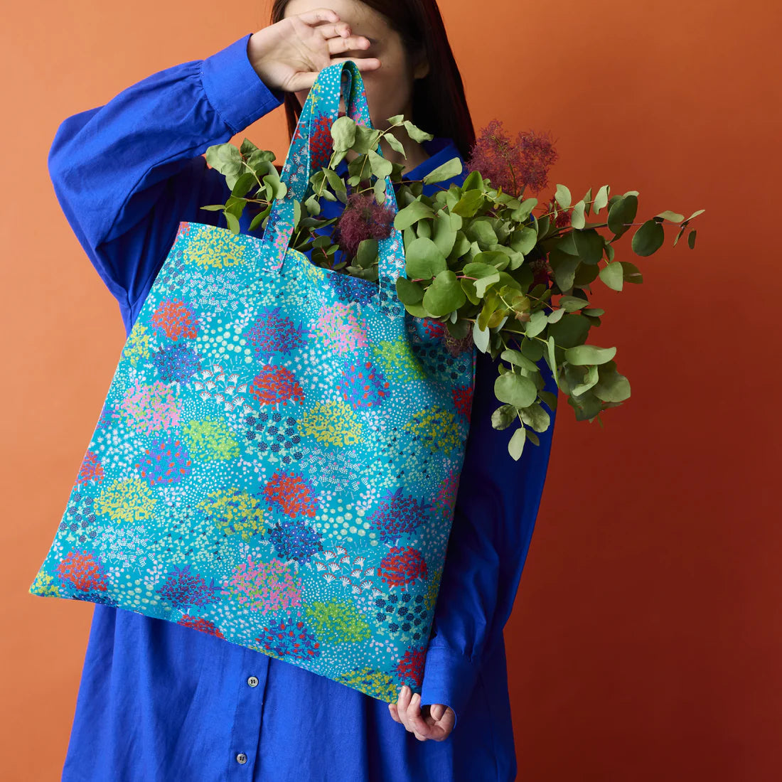 A person in a blue dress holds a colorful tote bag filled with greenery, including eucalyptus and other plants, against an orange background.