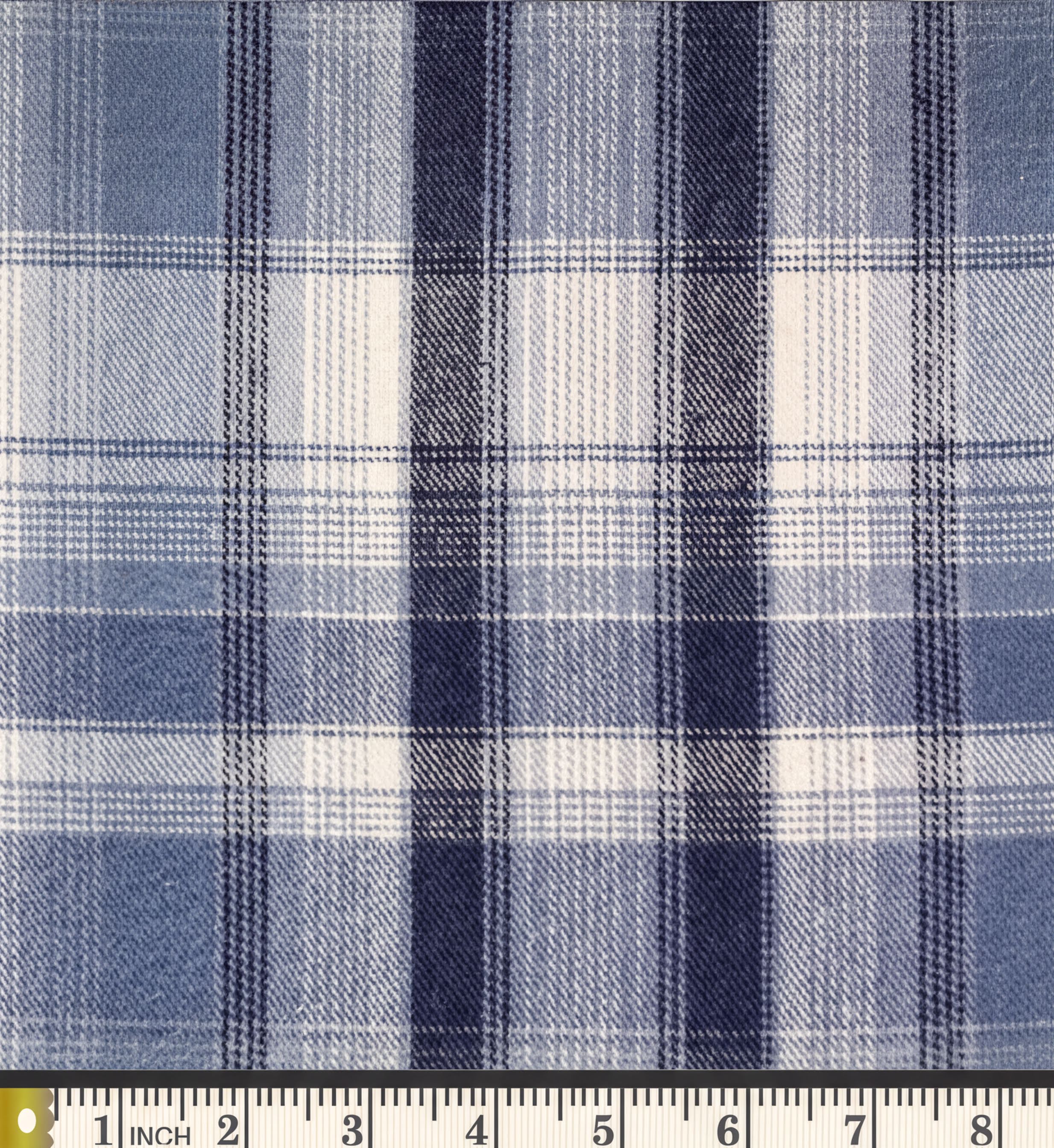 a close-up view of a fabric with a blue and white plaid pattern. The fabric appears to be a type of twill material, which is characterized by its diagonal lines and a repeating pattern of vertical and horizontal bands. The fabric is displayed on a ruler for scale, indicating its size.