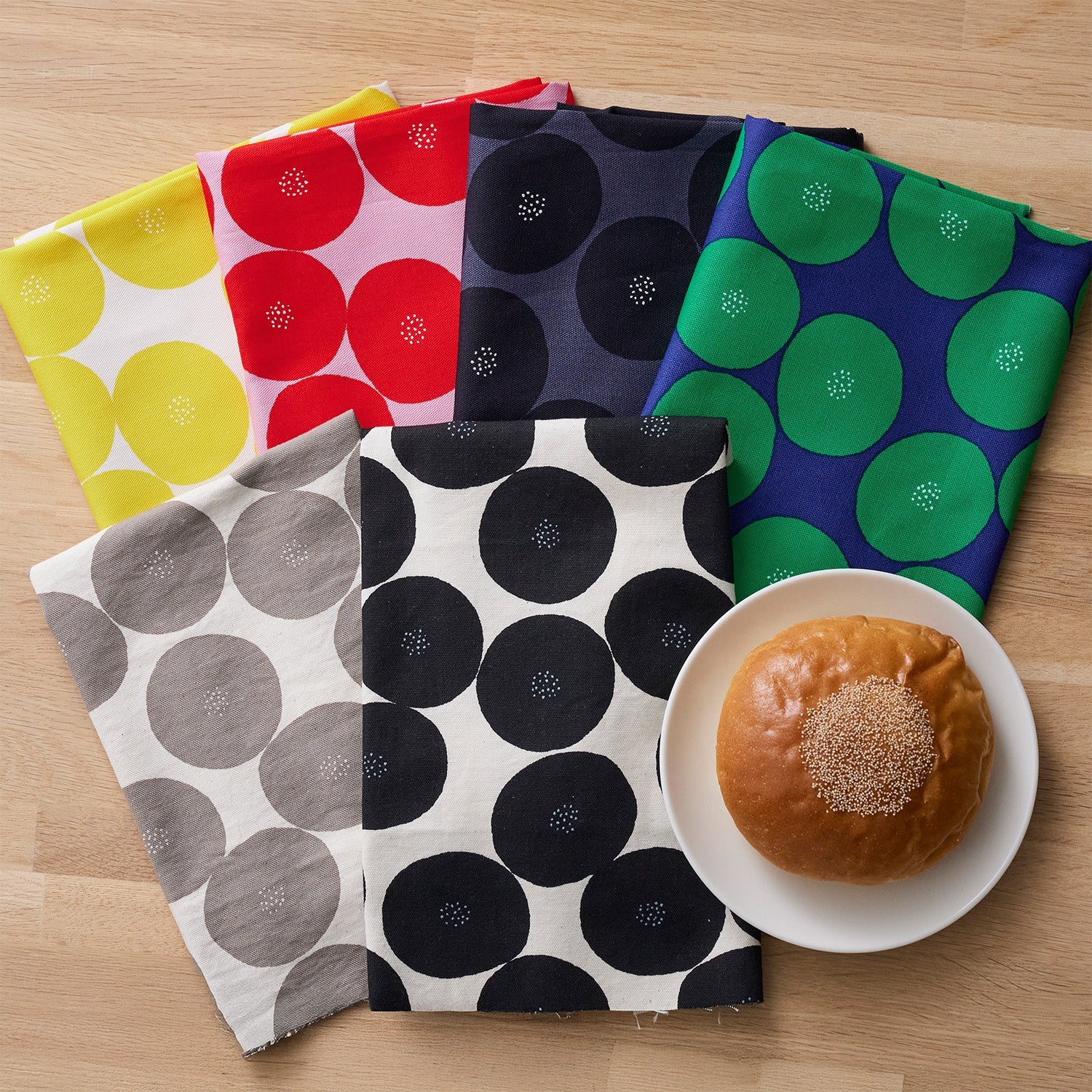 a variety of colorful fabric napkins arranged on a wooden surface, with a plate containing a pastry, likely a donut, in the foreground.