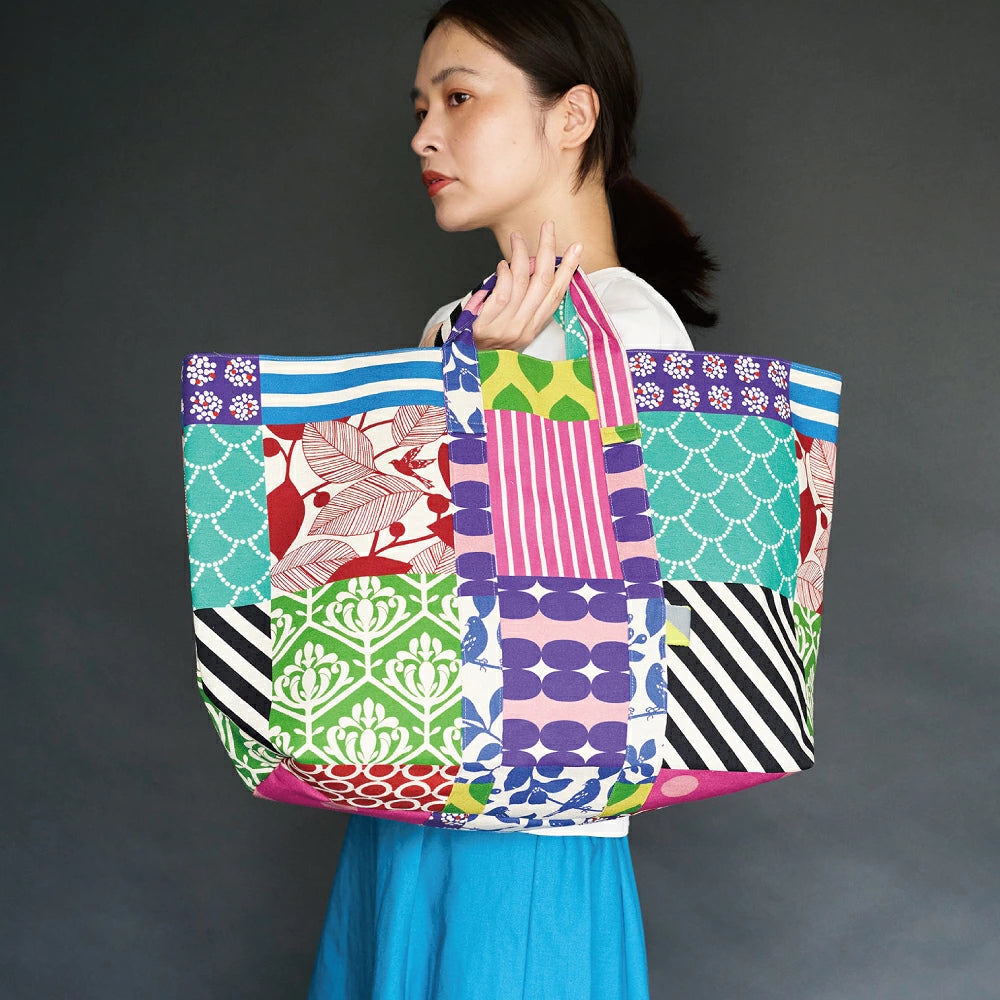 A woman with dark hair is holding a large, colorful tote bag with a variety of patterns and designs.