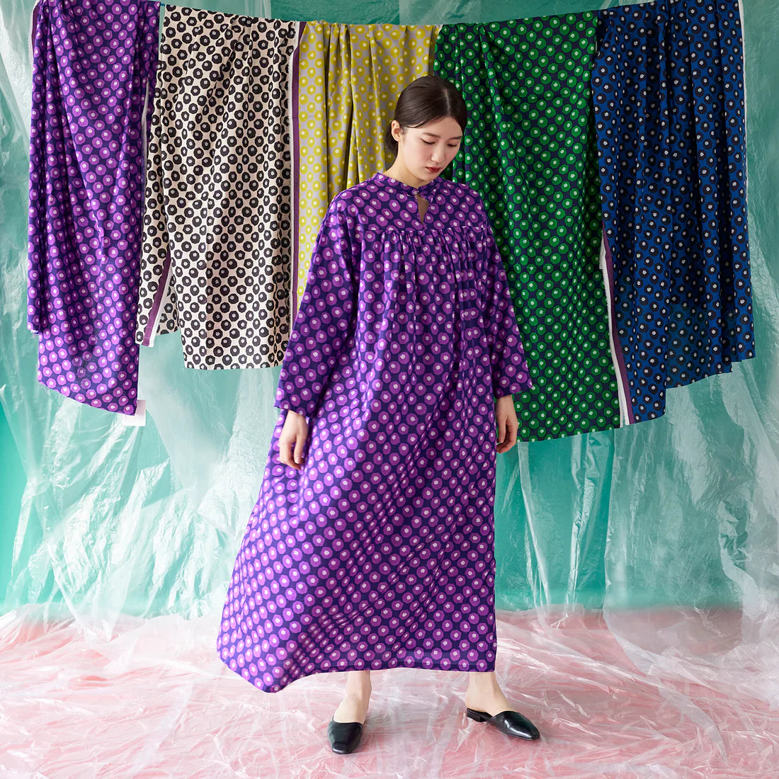 A woman wearing a long purple dress with a polka dot pattern stands in front of a backdrop with colorful dresses hanging on it.