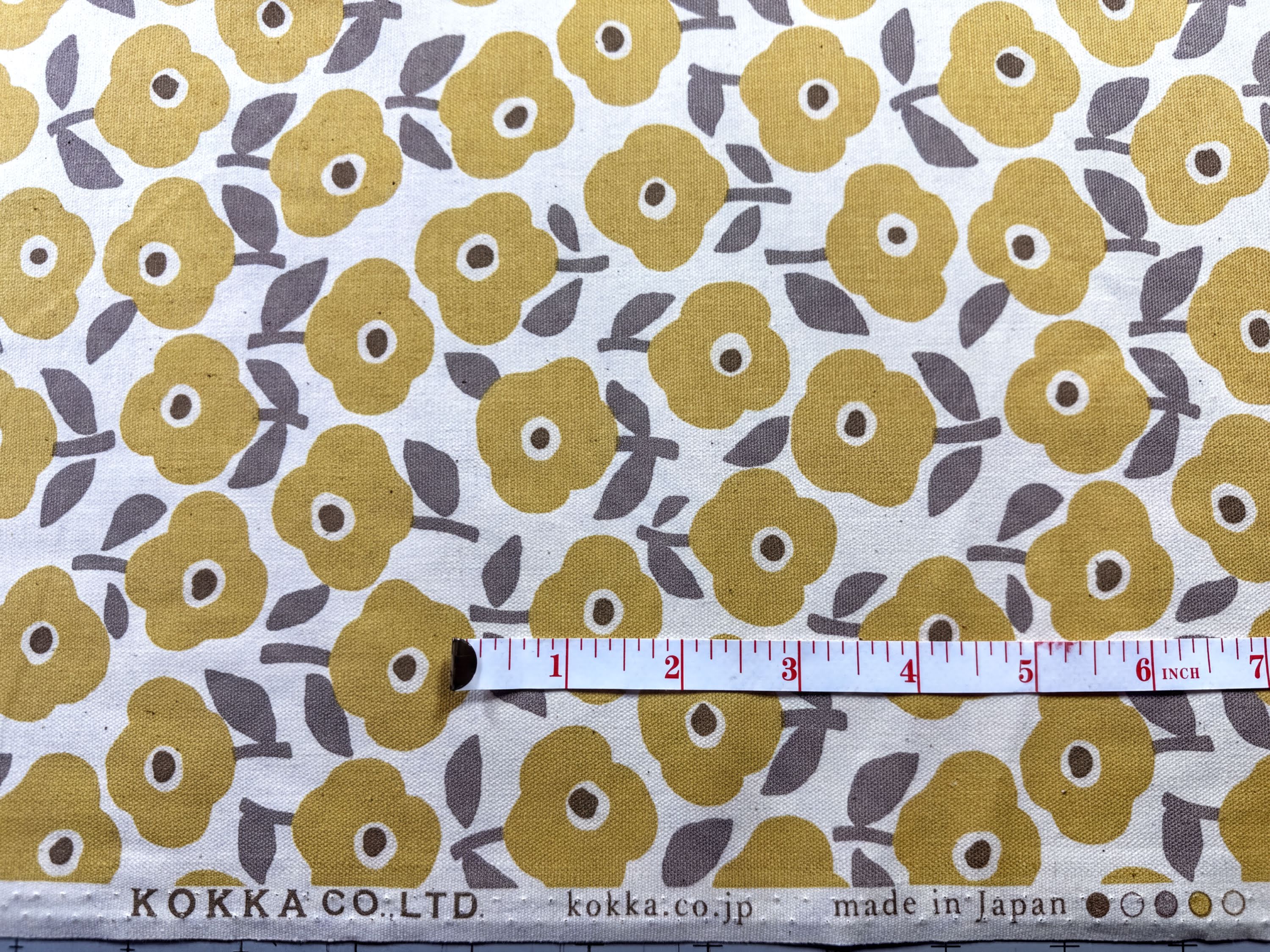 a fabric with a repeating pattern of yellow flowers on a white background. A ruler is placed in the center of the fabric, indicating the size of the fabric.
