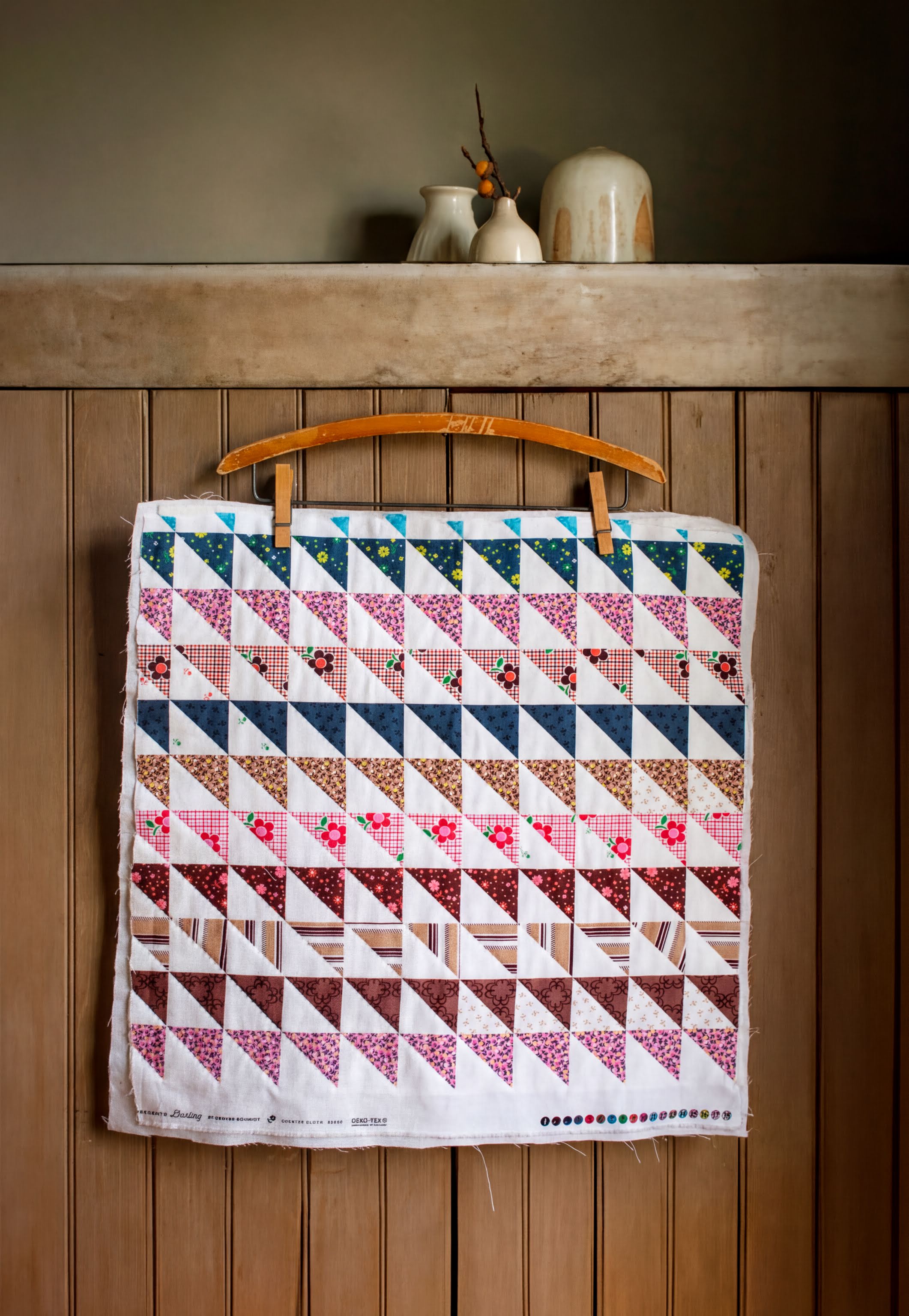 A colorful quilt with a geometric pattern is hanging on a wooden hanger on a wall.