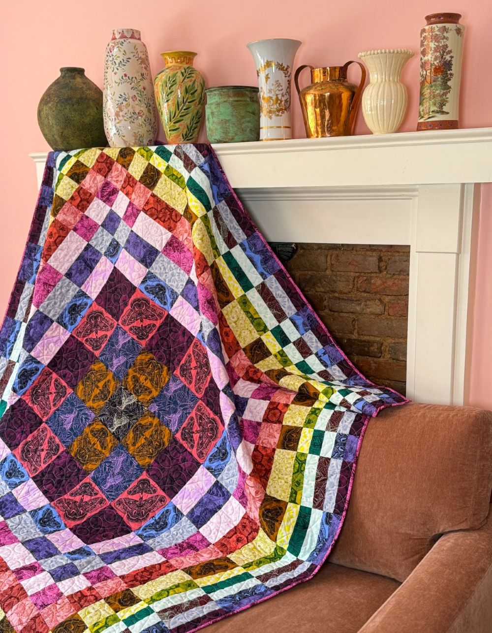 a vibrant, colorful quilt draped over a brown couch, with a variety of vases displayed on a shelf above it.