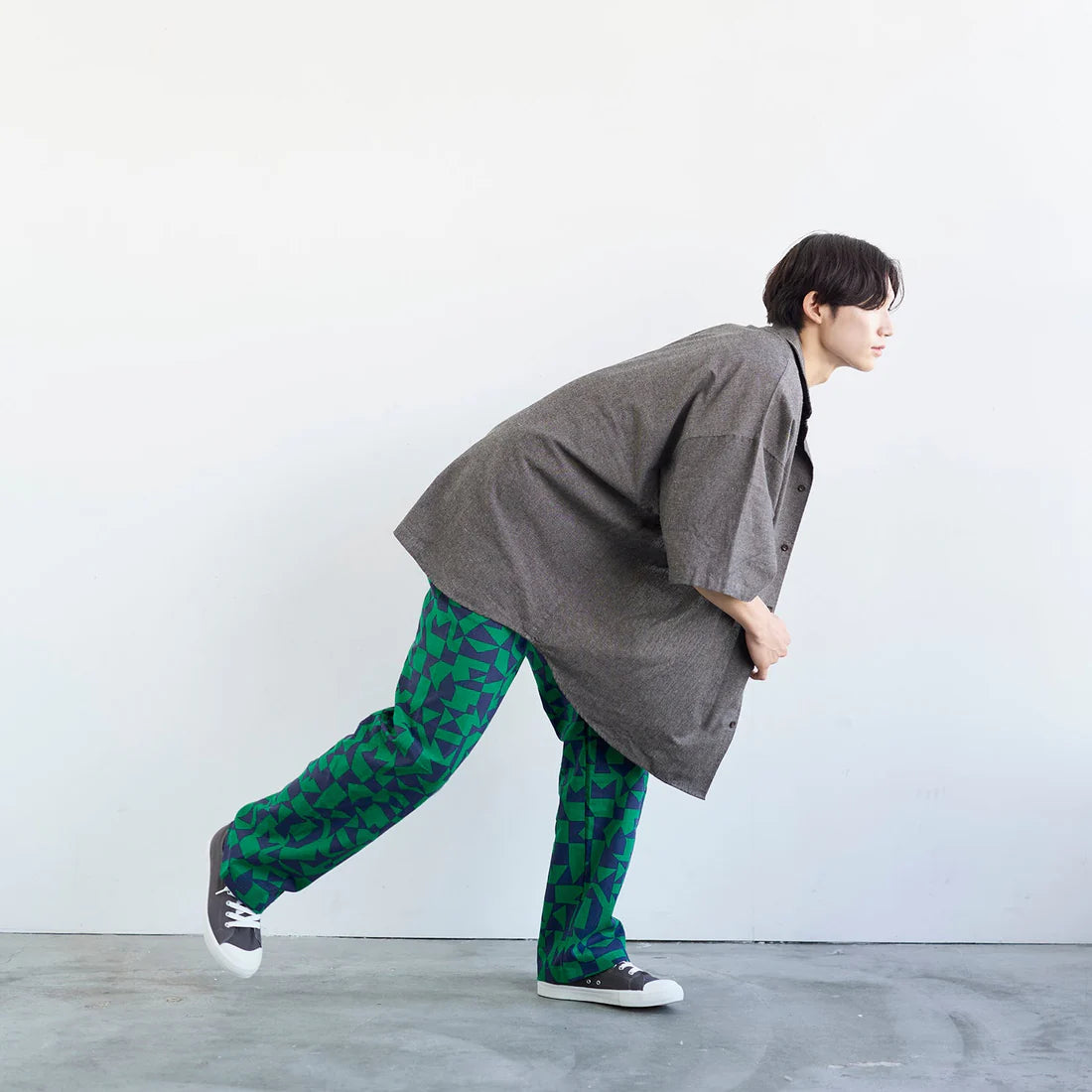 A person wearing a gray coat and green patterned pants is standing in a studio setting, with a neutral expression on their face.