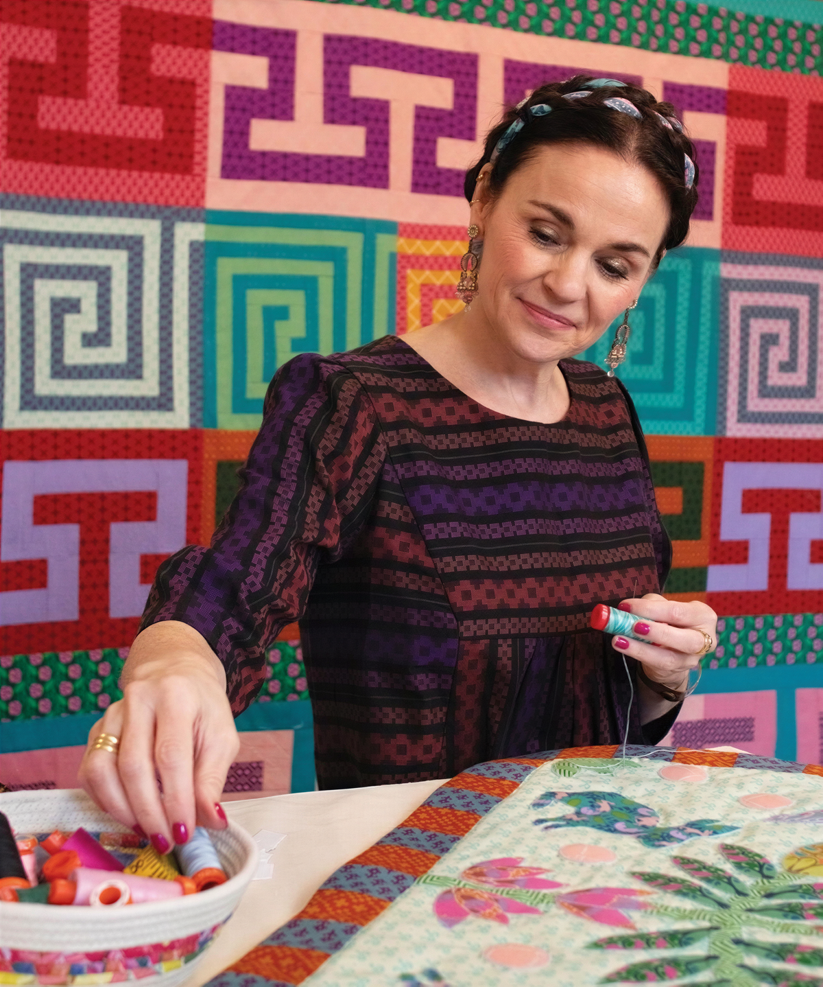 A woman is sitting at a table, working on a sewing project, with a colorful quilt in the background.