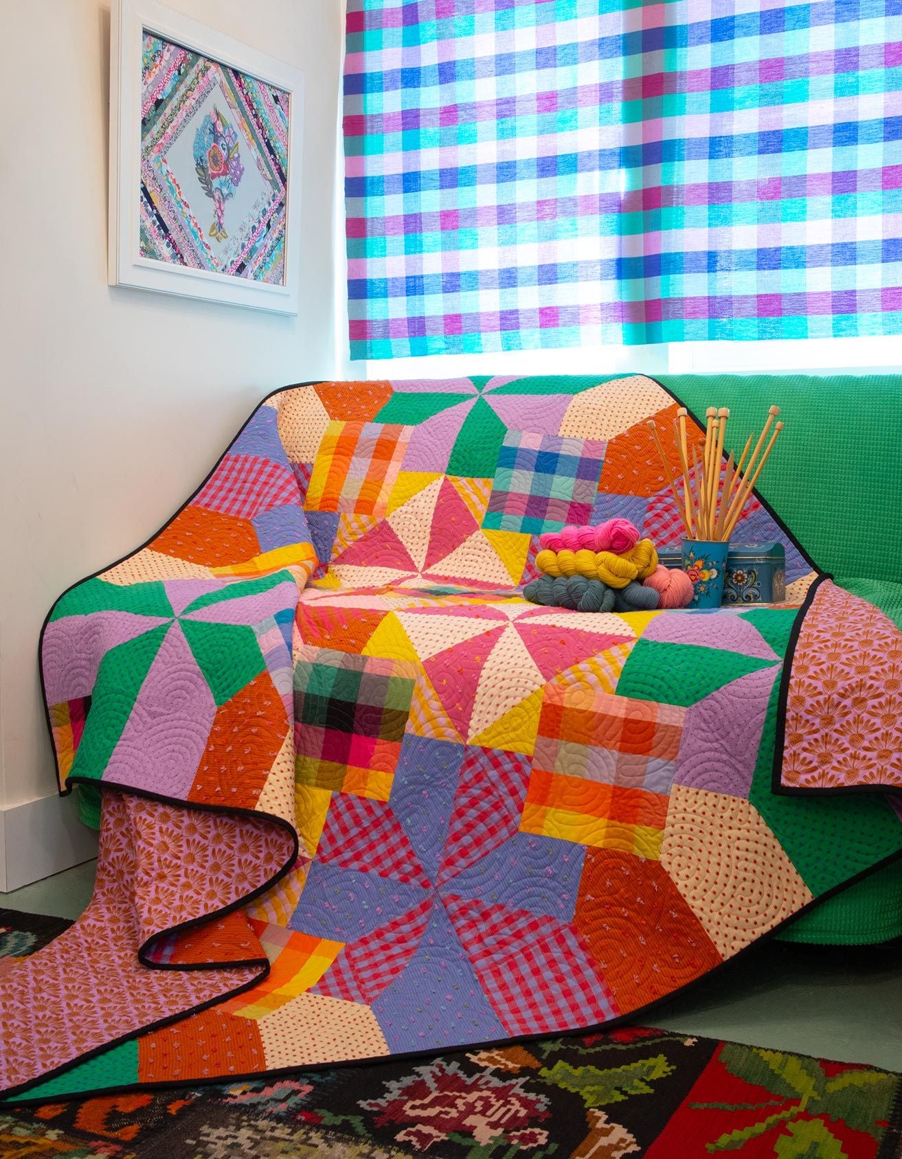 a vibrant, colorful quilt draped over a green couch, with a basket of knitting needles and balls of yarn nearby. The quilt is a patchwork of various geometric patterns in shades of pink, orange, green, and blue. The scene is set against a backdrop of a checkered curtain and a window, creating a cozy and inviting atmosphere.