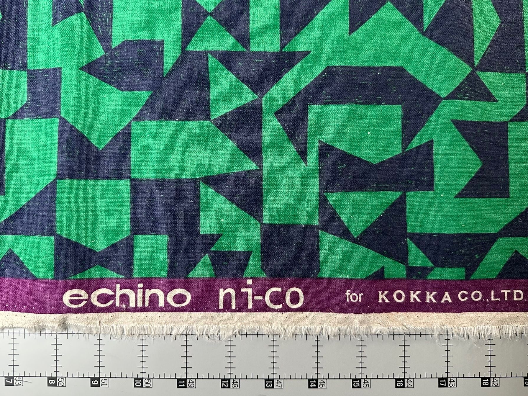 a fabric with a geometric pattern in shades of green and blue, with the text "echino ni-co" printed on it. The fabric is displayed on a ruler for size reference.