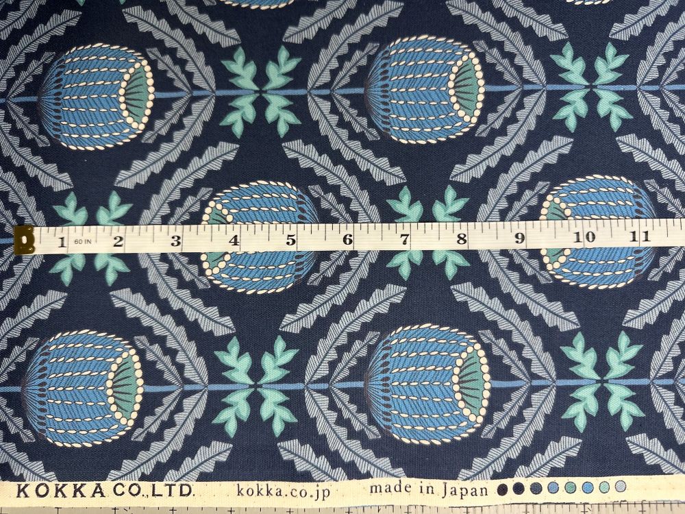 a fabric with a repeating pattern of blue and green flowers and leaves. The fabric is laid out on a table, and a ruler is placed next to it for scale.