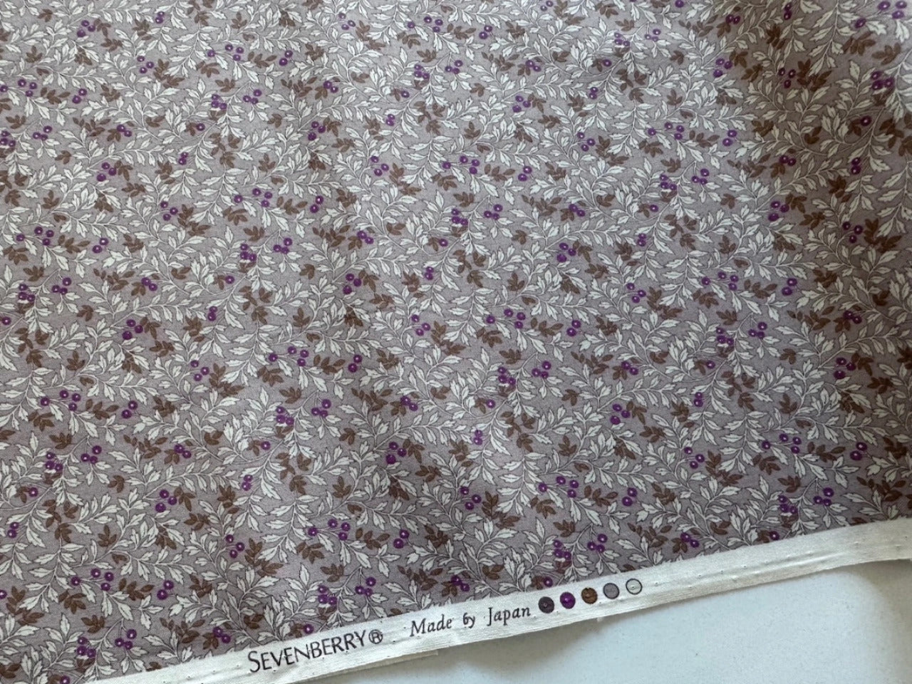 a fabric with a floral pattern in shades of gray and brown, featuring small purple flowers and leaves. The fabric appears to be made of a material with a floral print, and there is a label at the bottom of the fabric that reads "SEVENBERRY Made by Japan".