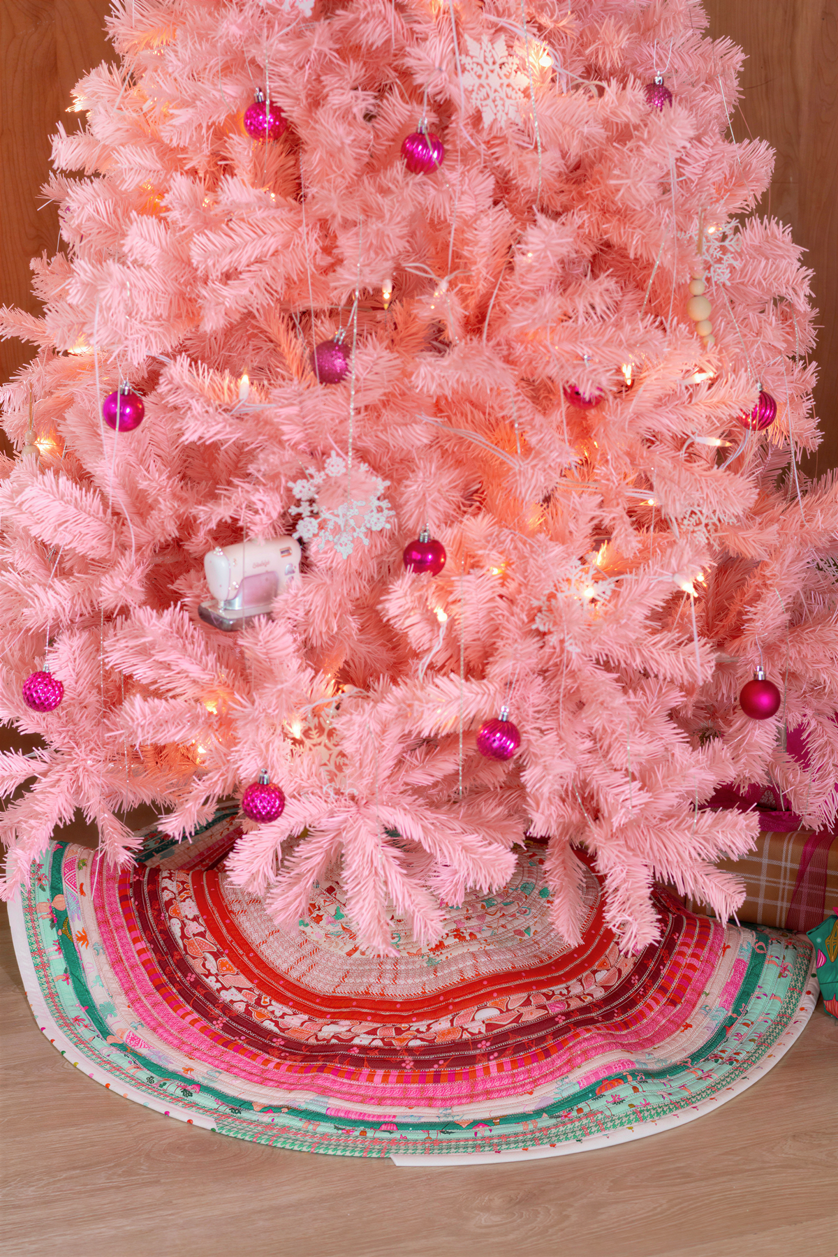 A pink Christmas tree adorned with ornaments and lights, surrounded by a colorful tree skirt.