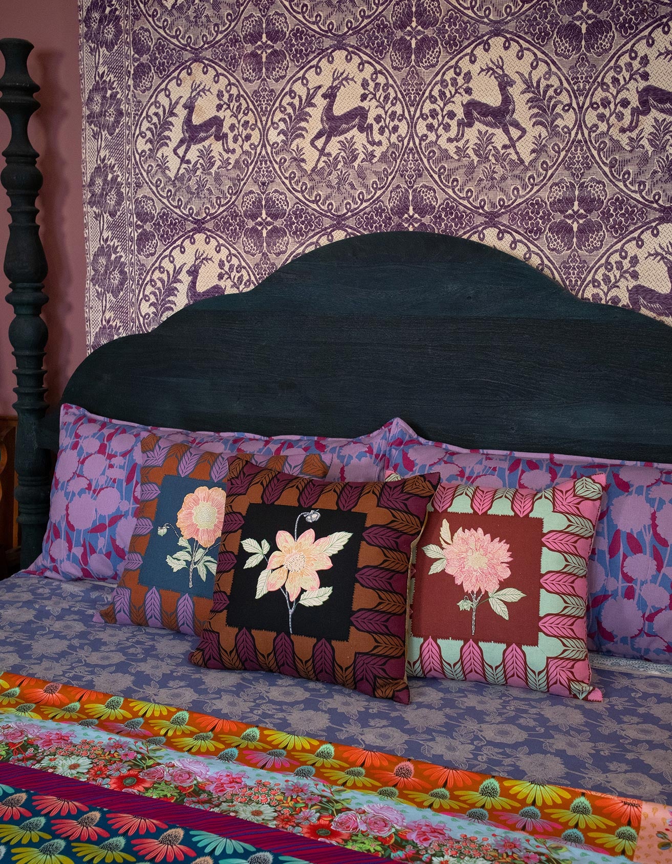 a bed with a black headboard and colorful pillows, including a floral patterned pillow and a pillow with a pink flower design. The bed is set against a purple and white patterned wallpaper.