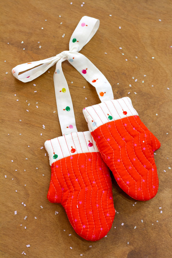 A pair of red and white patterned oven mitts with a white ribbon tied around them, placed on a wooden surface with scattered white specks.