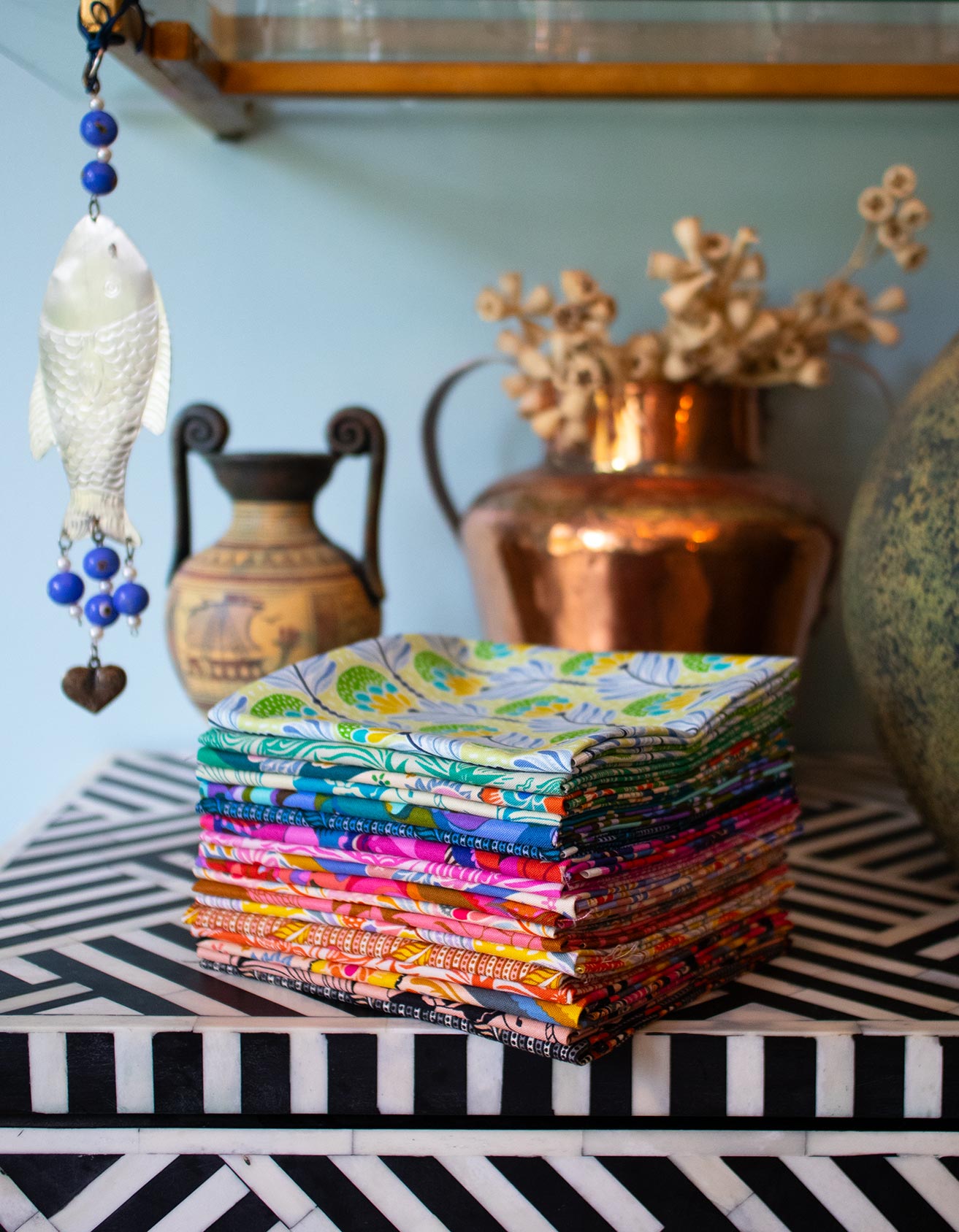 A stack of colorful fabric pieces is displayed on a black and white checkered surface, with a copper vase and blue and white fish ornament in the background.