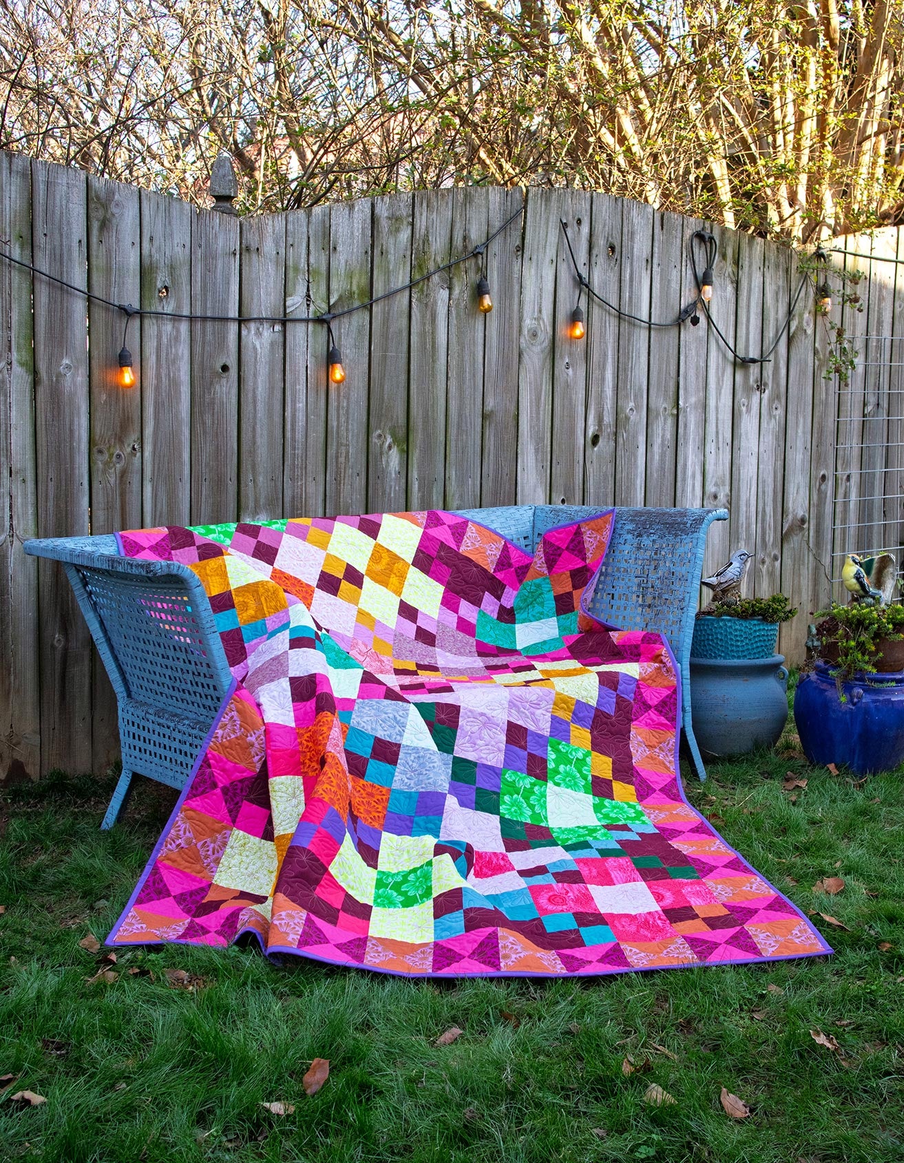 A vibrant, colorful quilt is spread out on a wicker chair in a backyard, with a wooden fence and string lights in the background.